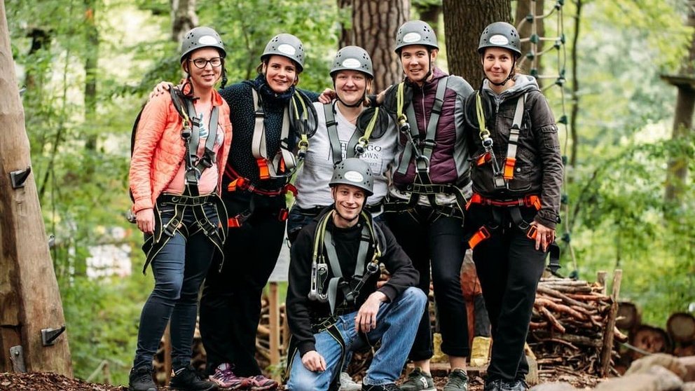 Gruppenfoto im Kletterpark: Sechs Personen mit Kletterausrüstung und Helmen posieren vor einem grünen Wald in Bad Saarow.