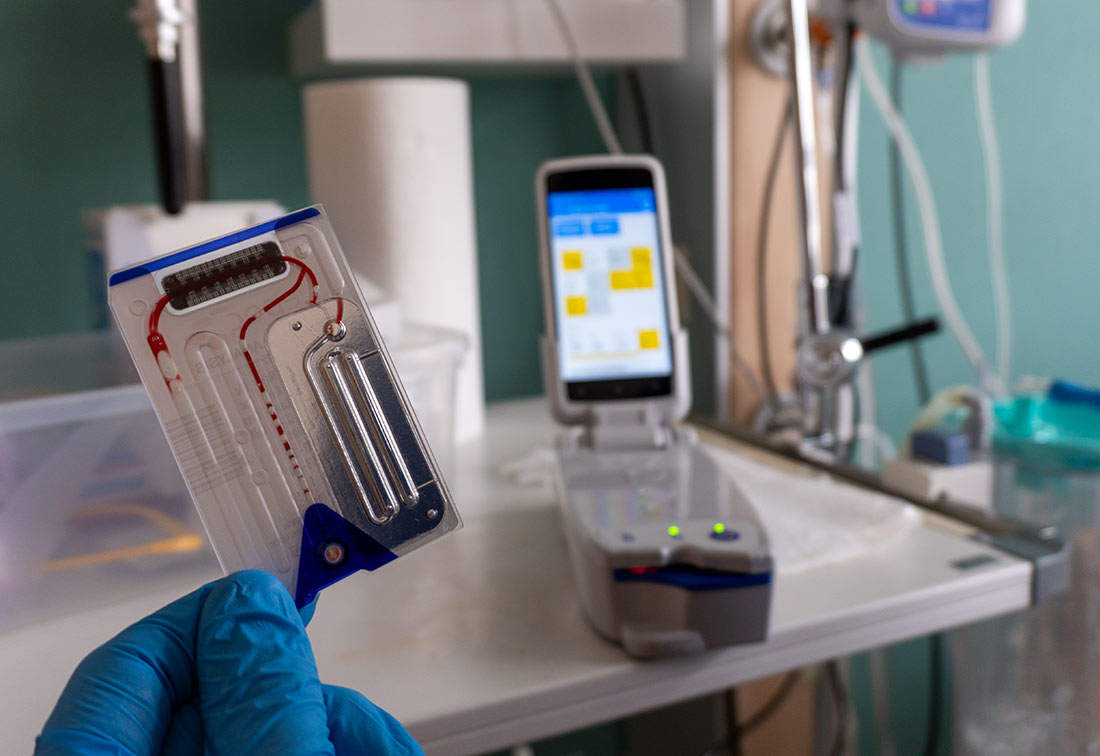 Eine Pflegefachkraft mit blauem Handschuh hält eine Testkarte in der Hand. Im Hintergrund ist ein BGA-Gerät zu sehen.