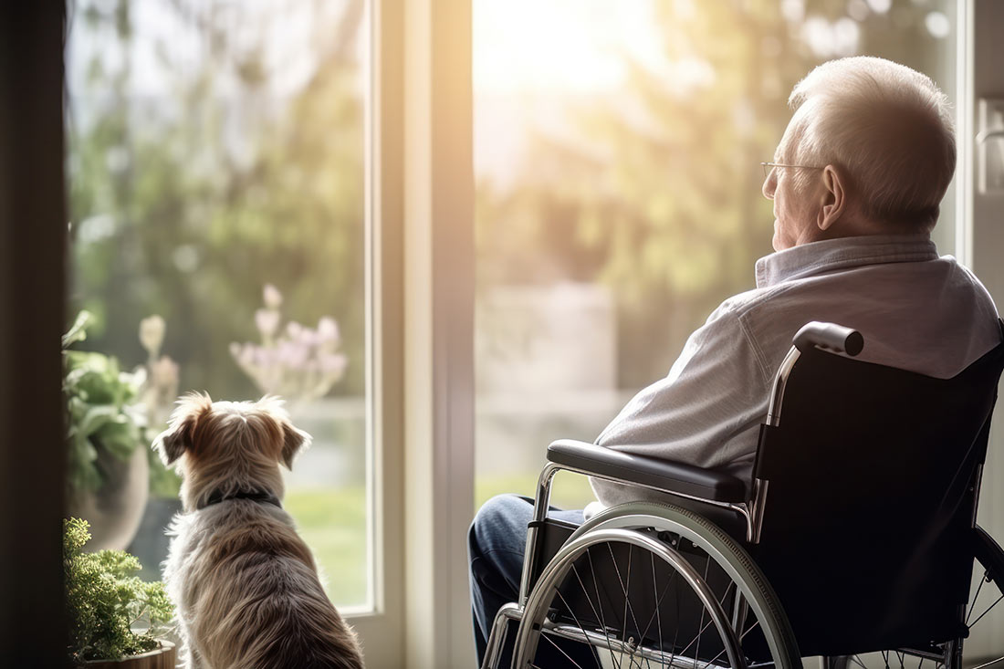 Ein Mann im Rollstuhl mit Hund zu Hause, der aus dem Fenster schaut.